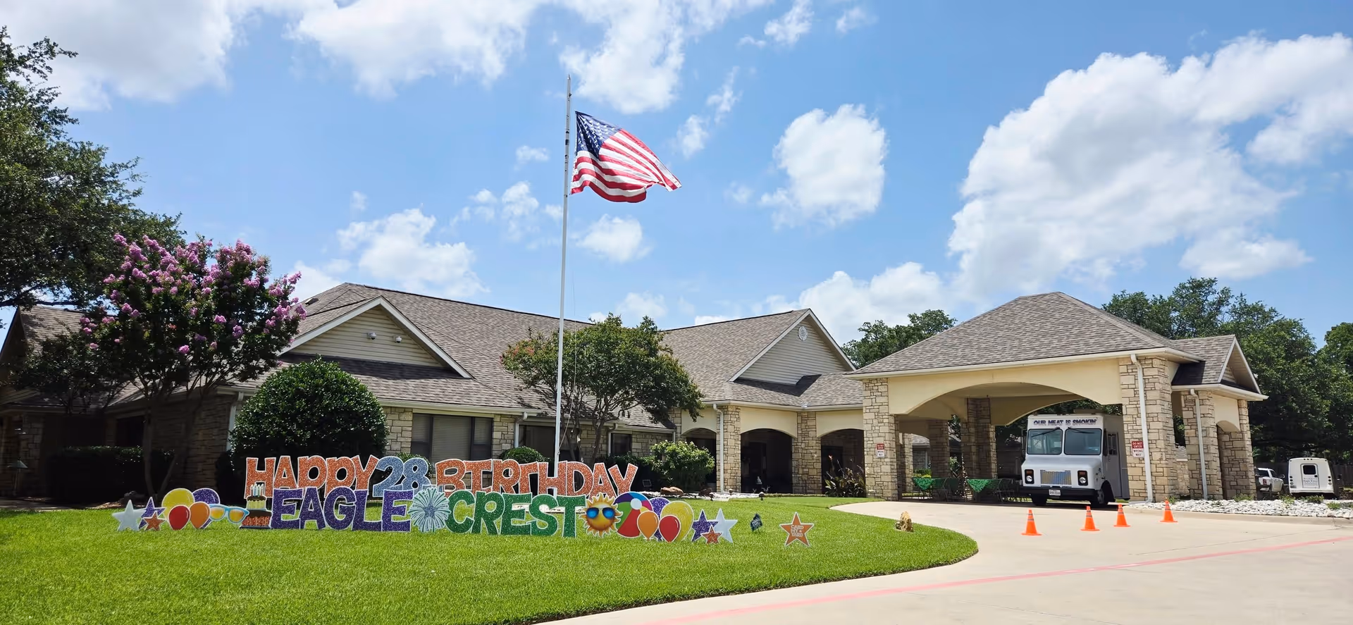 Exterior view of a single-story senior living facility building with a stone facade and a covered entrance. A tall flagpole with an American flag is centered on a well-maintained lawn. Colorful yard signs on the grass read 'Happy 28th Birthday Eagle Crest' with decorative balloons and stars. The sky is partly cloudy with blue patches.
