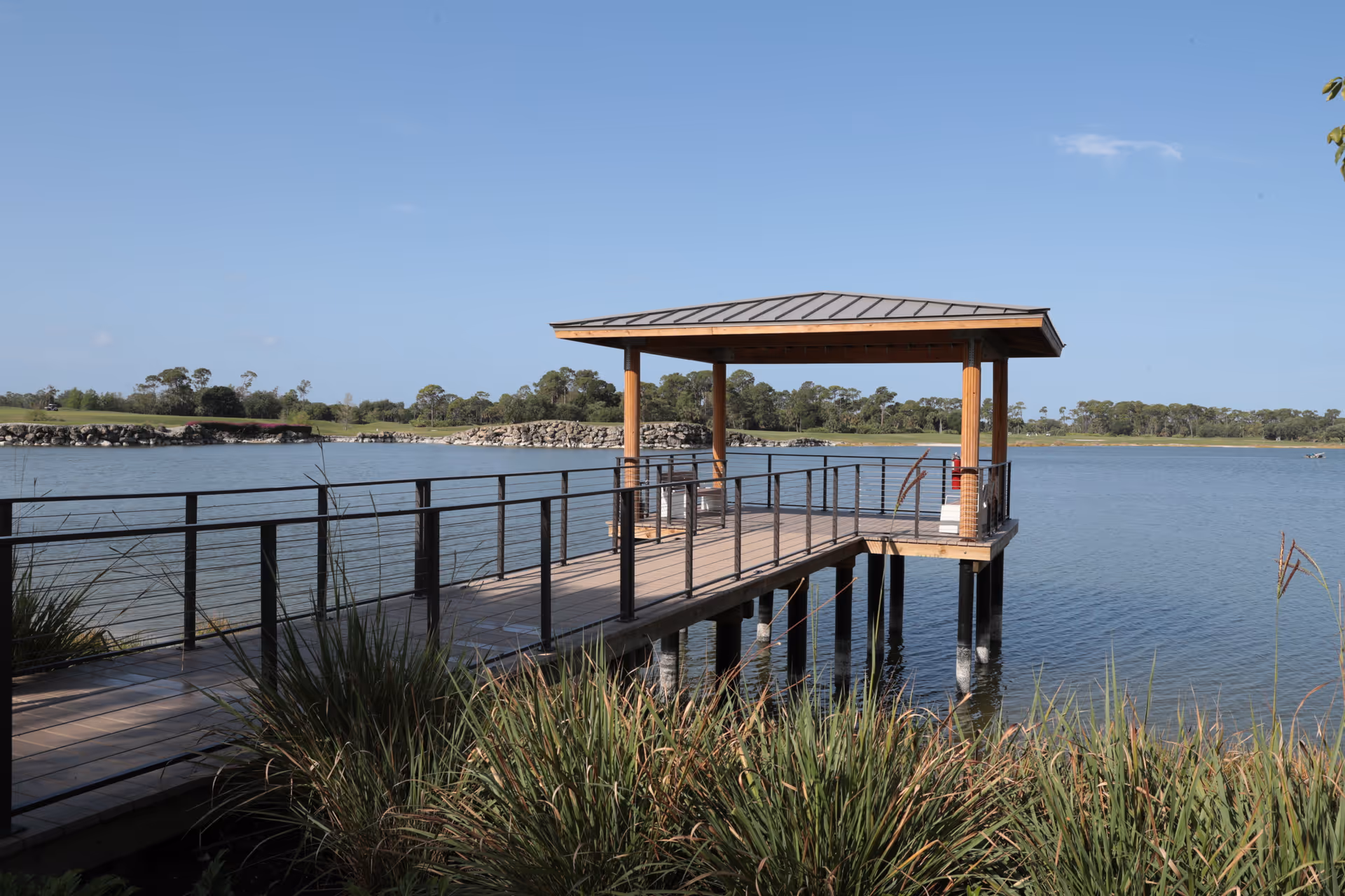 A wooden pier with a covered gazebo extends over a calm lake surrounded by greenery and trees under a clear blue sky.