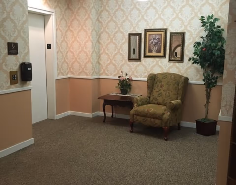 A cozy corner inside a facility with a patterned armchair, a small wooden table with a flower arrangement, three framed wall decorations including two mirrors and a floral painting, a potted plant, and an elevator door marked with the number 5. The walls have a two-tone design with wallpaper on the upper half and peach paint on the lower half.