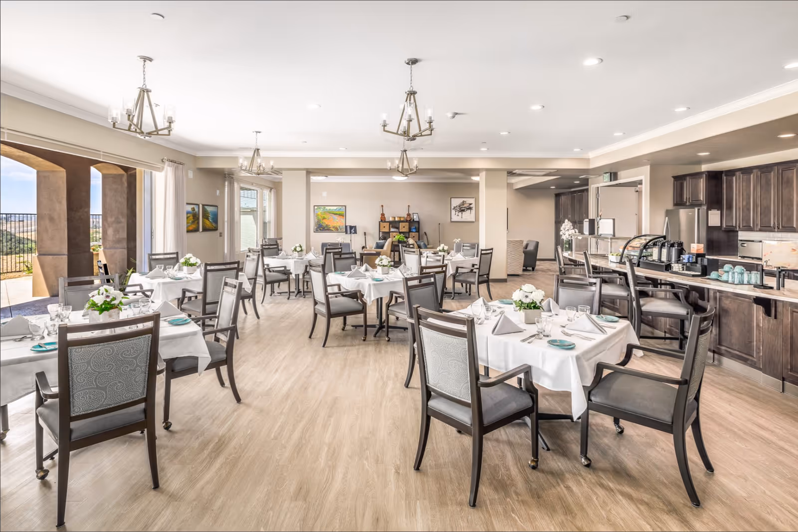 Bright, spacious dining room with multiple set tables and chairs, chandeliers, and a serving counter.
