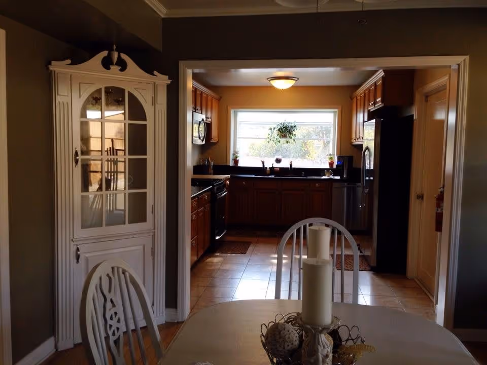 View from a dining area into a kitchen. The dining area has a white table with a decorative centerpiece and white chairs. The kitchen features wooden cabinets, a window with a hanging plant, a stove, microwave, dishwasher, and refrigerator. The walls are painted in warm tones and the floor is tiled.