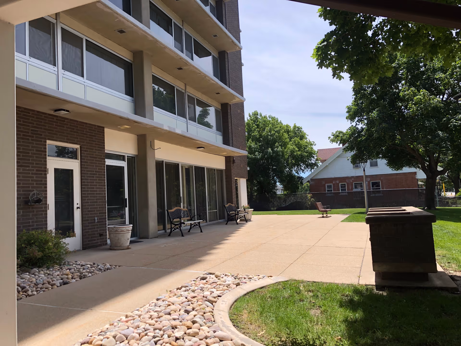 Outdoor patio area of Fellowship Manor with benches, a paved walkway, green grass, trees, and a brick building in the background under a clear sky.