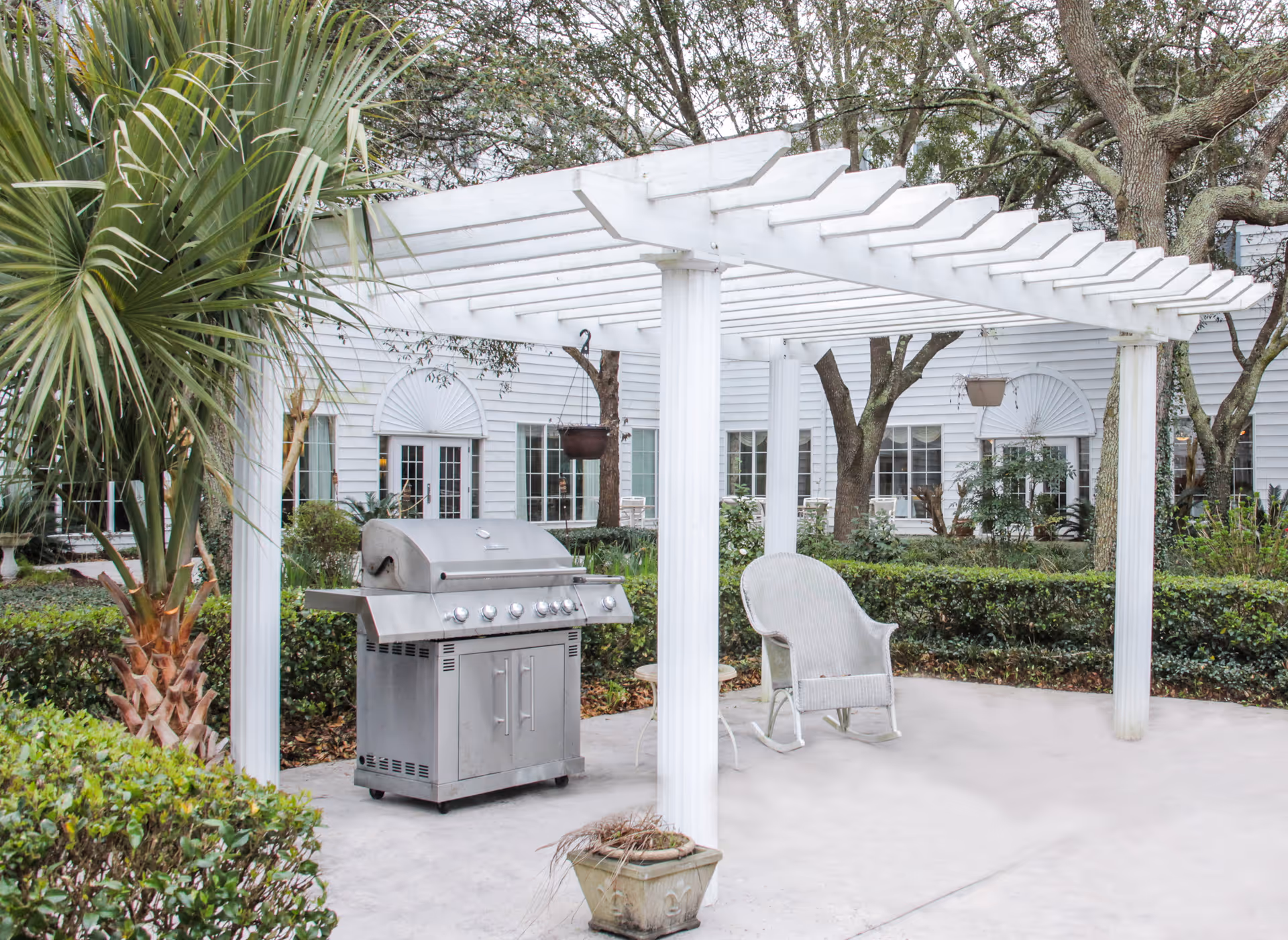 Outdoor patio area with a white pergola, a stainless steel barbecue grill, a white wicker rocking chair, and surrounding greenery including palm trees and bushes. In the background, there is a white building with multiple windows and doors.