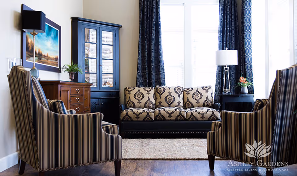 A cozy living room with a patterned beige and black sofa centered against a window with dark blue curtains. Two striped armchairs face the sofa. There is a wooden cabinet with glass doors and a chest of drawers with a lamp and a plant on top. A framed landscape painting hangs on the wall.
