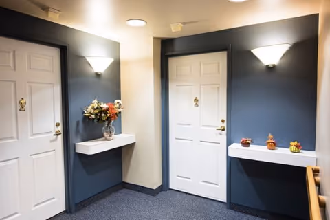 Carpeted interior hallway with two white apartment doors in dark blue alcoves, wall sconces, floating shelves and decorative vases.