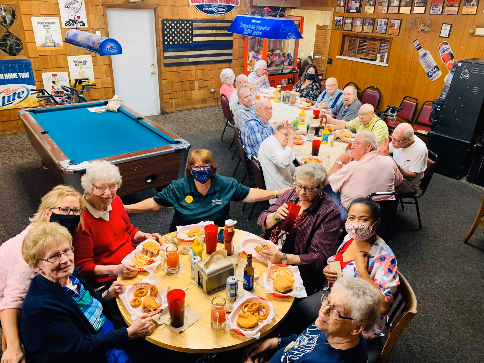 A group of elderly people and caregivers sitting around two tables in a recreational room, enjoying a meal with burgers, fries, and drinks. The room has wood-paneled walls, a pool table, and various posters and decorations including a black and white American flag and beer advertisements. Some individuals are wearing face masks.