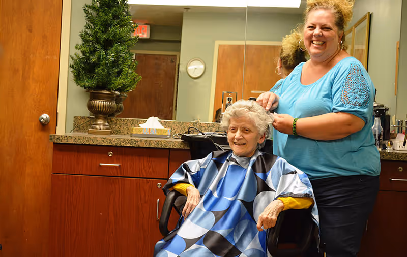 An elderly woman sitting in a salon chair wearing a blue and black patterned cape while a smiling hairstylist stands behind her, styling her hair. The setting appears to be a hair salon area within a facility, with a large mirror, wooden cabinets, a small potted plant, and a tissue box on the countertop.