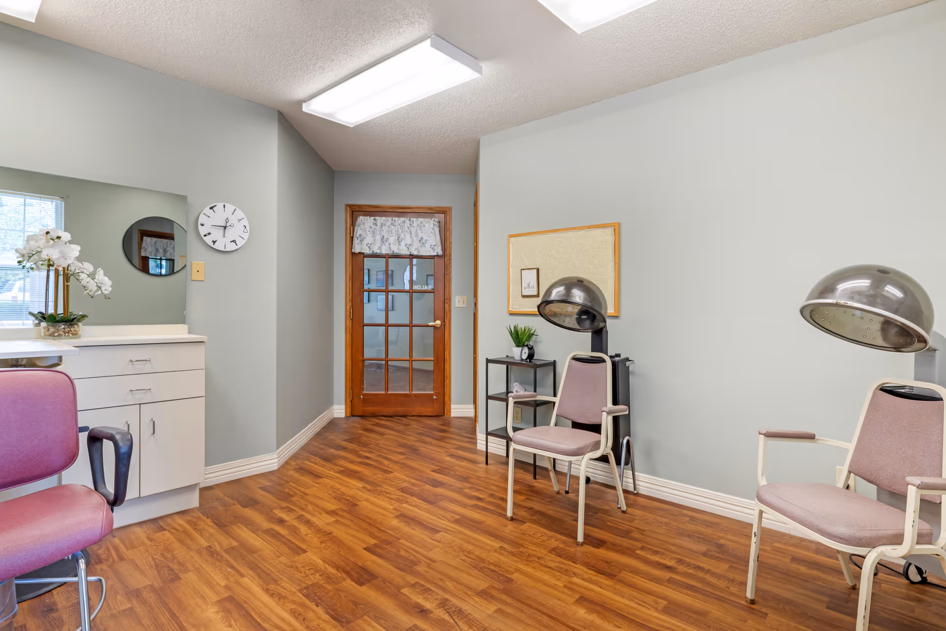 Small salon area with hooded hair dryers, padded chairs, a vanity counter, and a glass-paneled wooden door on wood flooring.