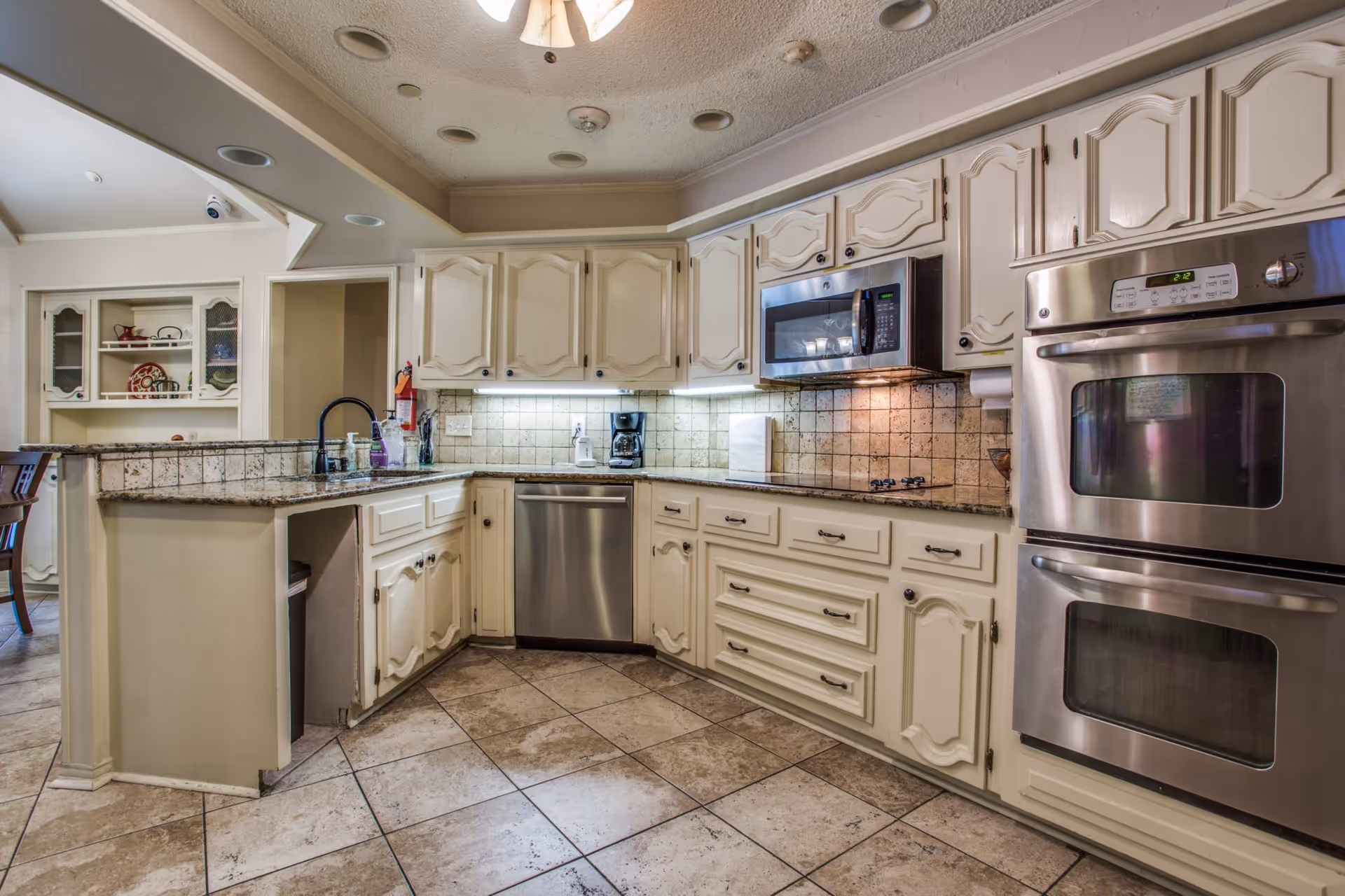 A spacious kitchen with cream-colored cabinets, granite countertops, and tiled floor. The kitchen features a built-in microwave, double oven, dishwasher, coffee maker, and a sink with a black faucet. There is a small open shelf area with decorative items visible in the background.