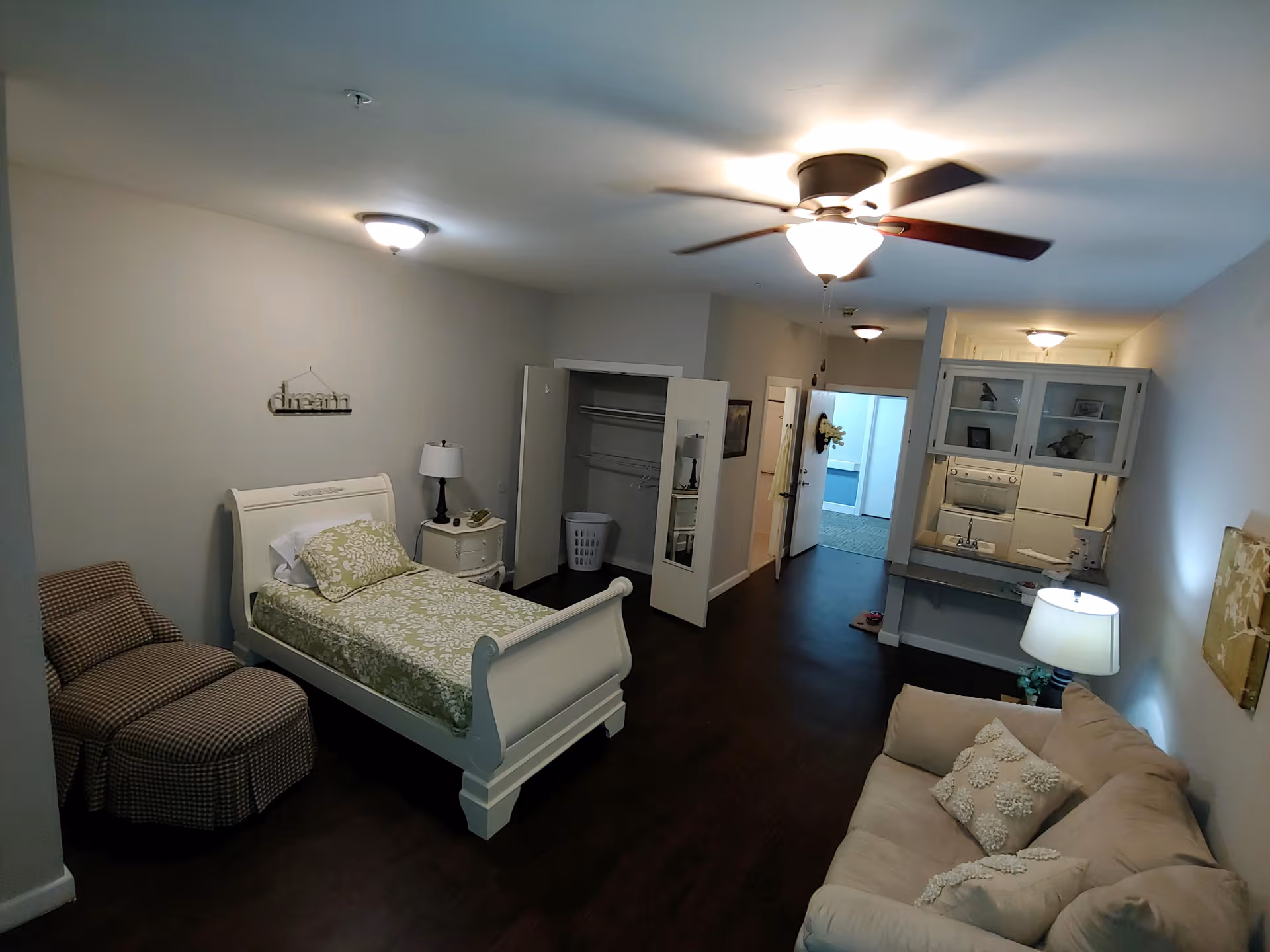 A senior living facility room with a single bed covered in a green and white patterned bedspread, a nightstand with a lamp, an open closet with a laundry basket inside, a beige sofa with decorative pillows, a ceiling fan with light, and a small kitchenette area with cabinets, a microwave, and a sink.