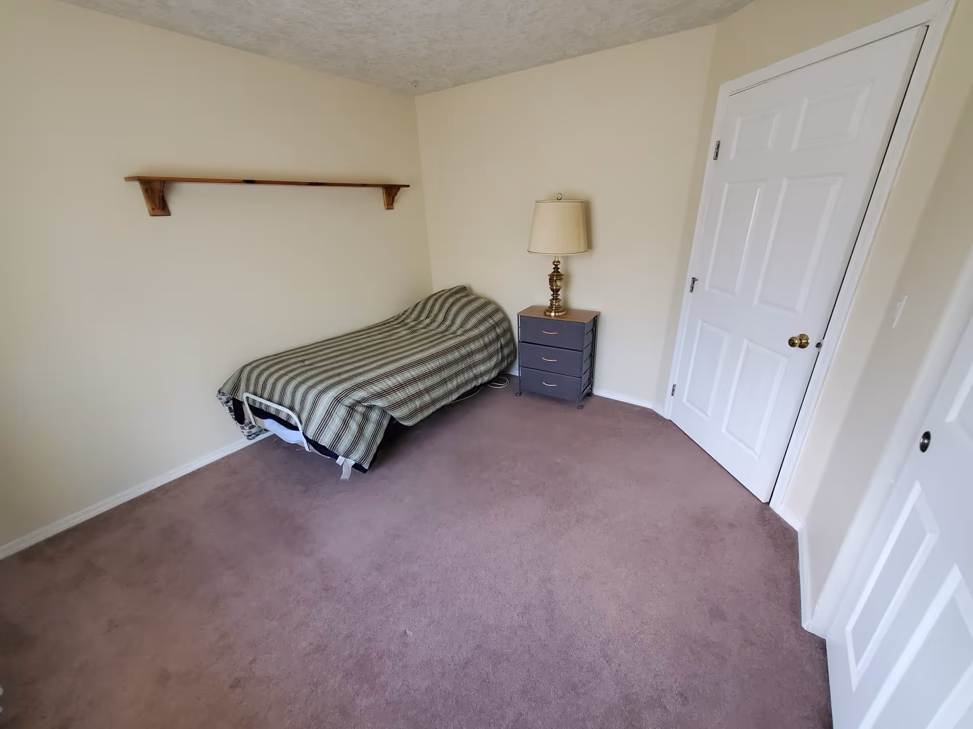 A small bedroom with a single bed covered in a striped blanket, a nightstand with a lamp, a wooden shelf mounted on the wall, and two white doors. The room has beige walls and carpeted floor.