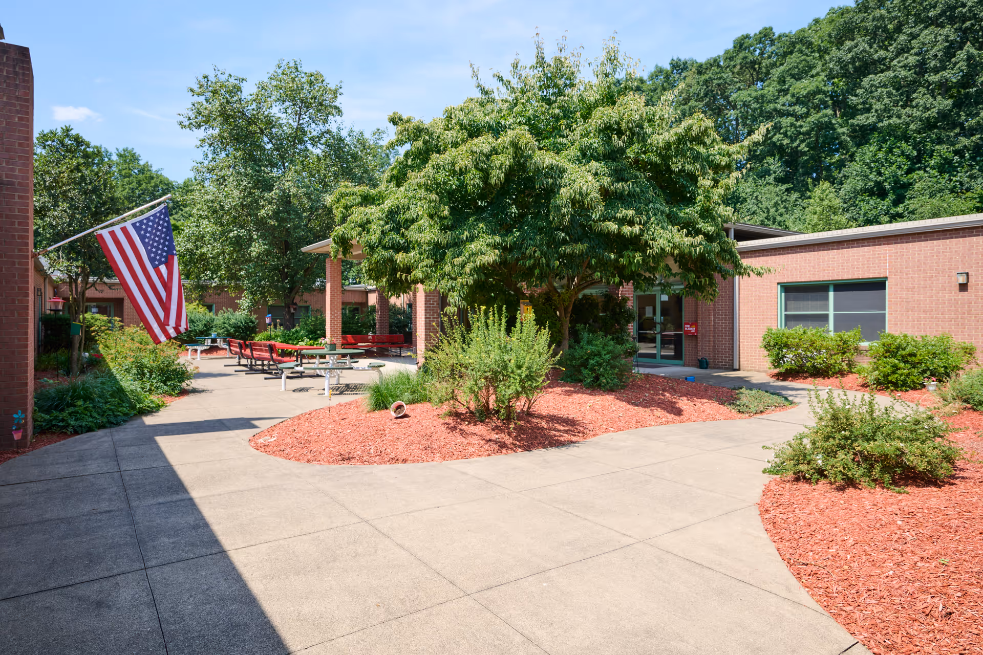Outdoor courtyard area of Mountain Top Rehabilitation and Healthcare Center featuring a paved walkway, landscaped garden beds with red mulch, green shrubs, and a large tree. There are several picnic tables and benches under a covered area. An American flag is displayed on the left side attached to the building. The building is made of brick and surrounded by trees and greenery.