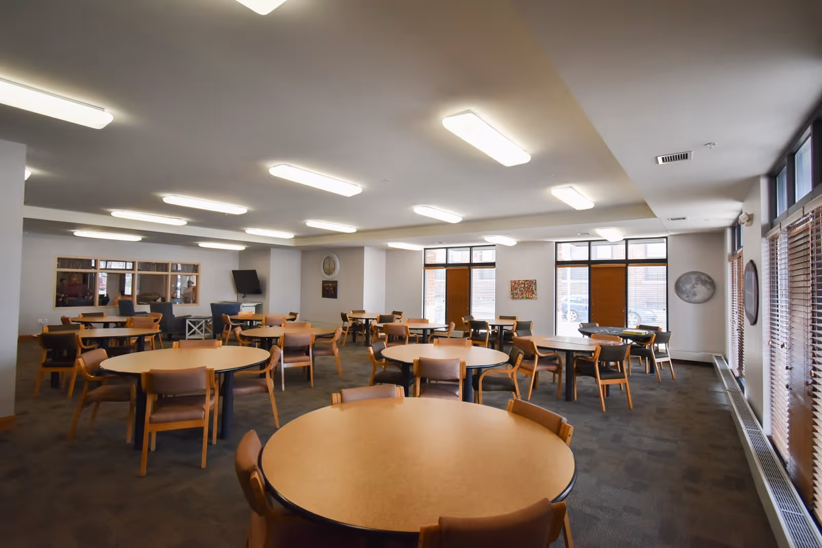 A spacious dining room in Lyons Court Senior Apartments with multiple round tables and wooden chairs arranged neatly. The room has large windows with blinds, carpeted flooring, and ceiling lights providing ample illumination. There is a television mounted on the wall and some artwork hanging on the walls.