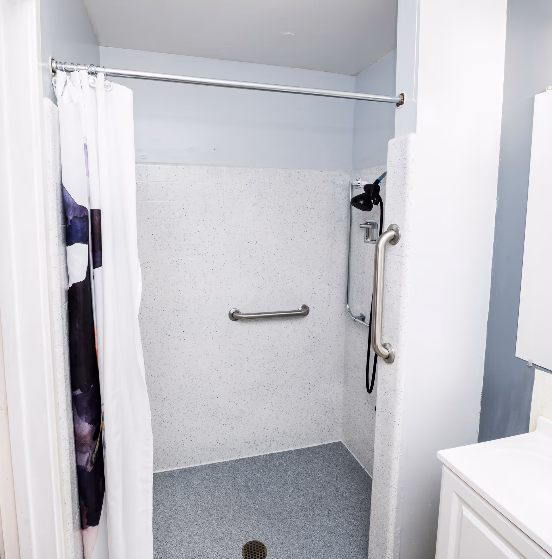 A clean, accessible shower area with a white and gray color scheme. The shower has a stainless steel grab bar on the wall and another grab bar on the side wall near the showerhead. There is a white shower curtain with a dark floral pattern partially drawn to the side. The floor is textured gray, and a white sink with a cabinet is partially visible on the right side.