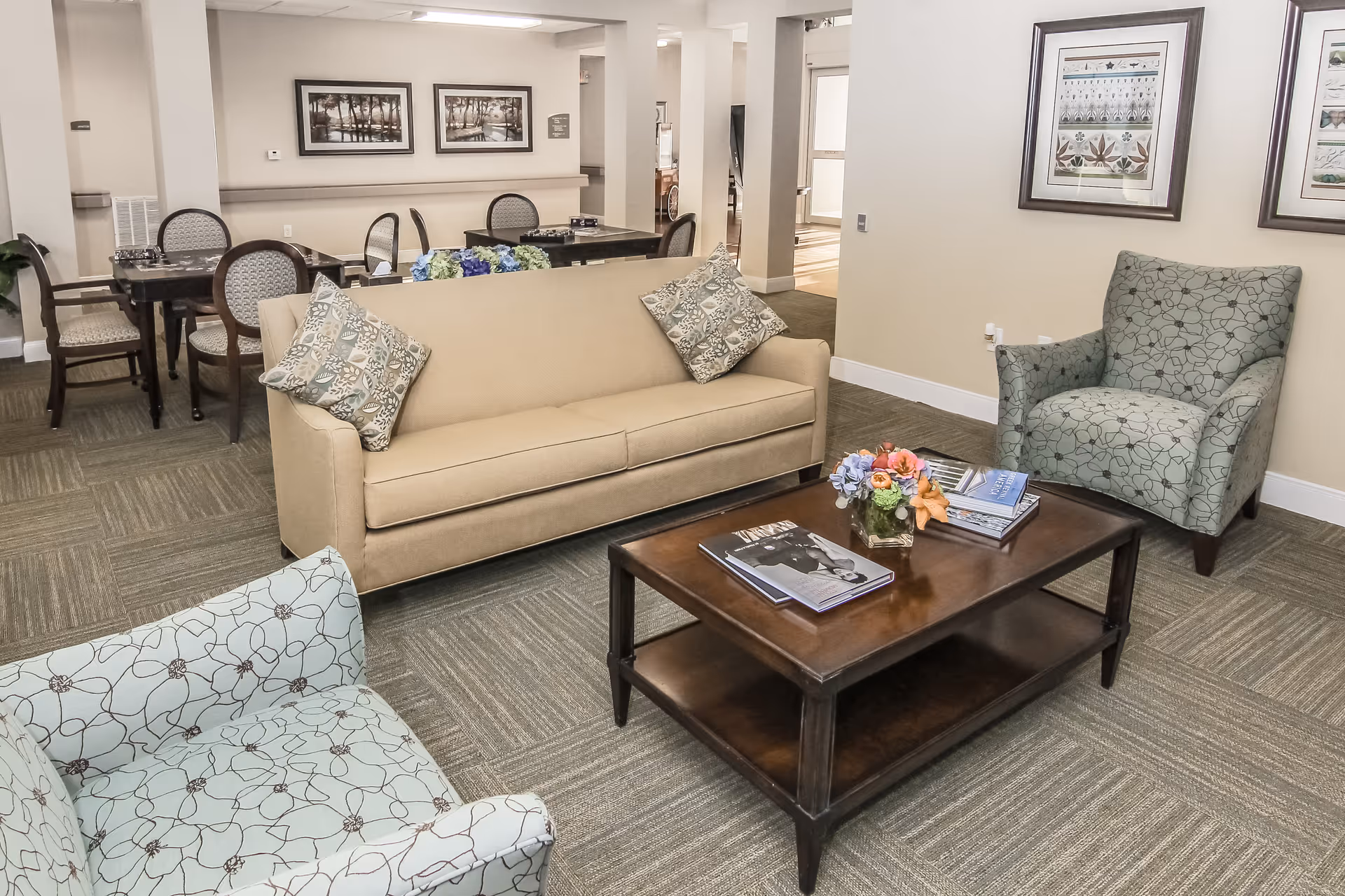 A cozy living room area with a beige sofa adorned with patterned pillows, two patterned armchairs, and a dark wooden coffee table with books and a small flower arrangement. In the background, there are tables and chairs arranged in a dining area, with framed artwork on the walls and neutral-colored carpeting.