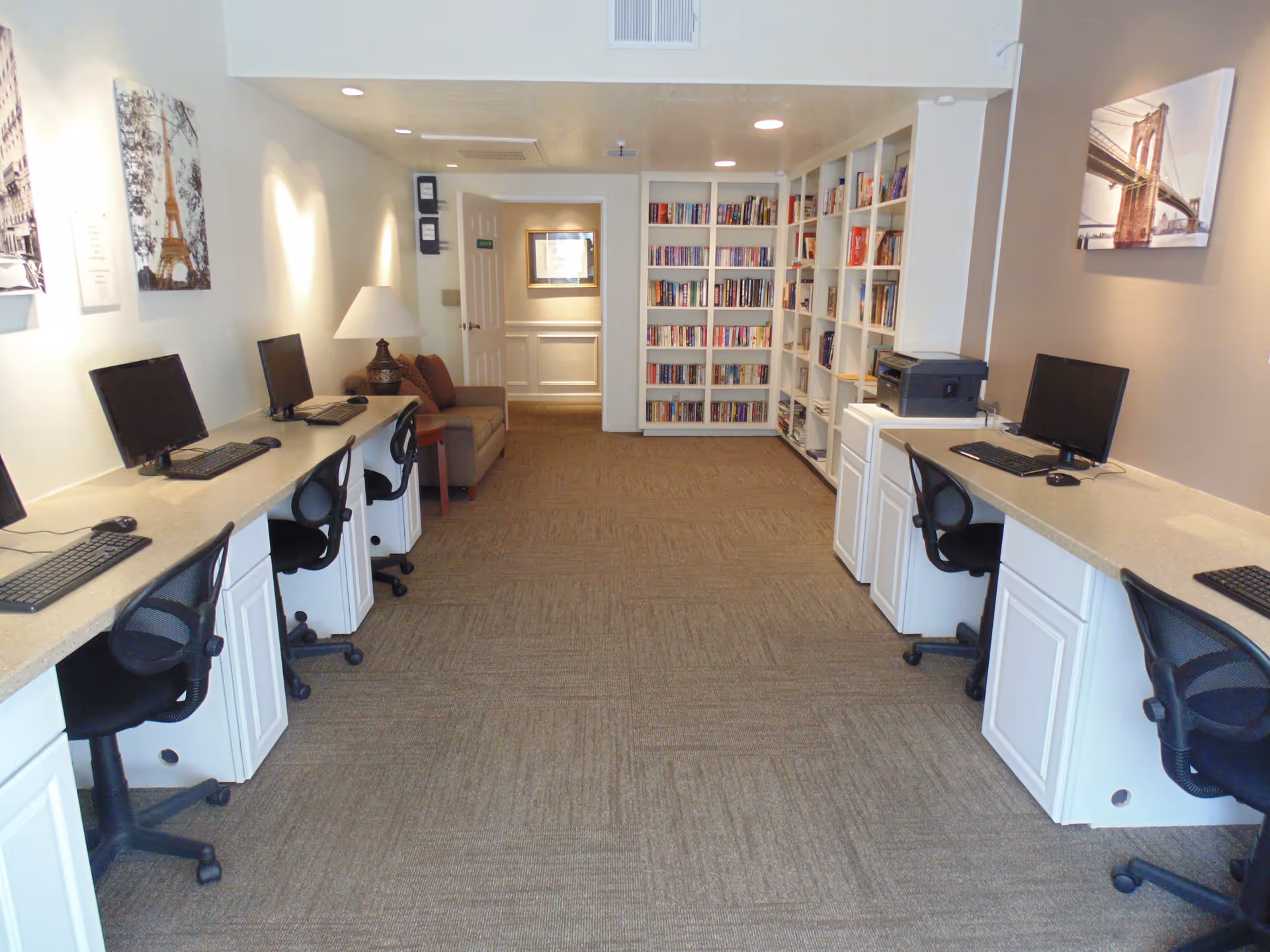 A computer room with multiple desktop computers and black office chairs arranged along both sides of the room. There is a beige carpeted floor, white walls with framed pictures, a small couch with a lamp on a side table, and a large bookshelf filled with books at the far end of the room.