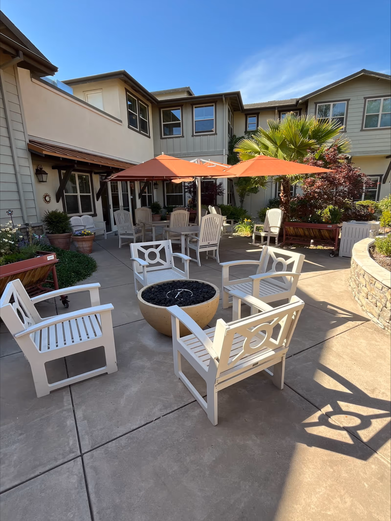 Outdoor patio area at CountryHouse At Granite Bay with white wooden chairs arranged around a circular fire pit. There are orange umbrellas providing shade over a table with additional chairs. The patio is surrounded by plants, including a palm tree and other greenery, with a two-story building in the background under a clear blue sky.