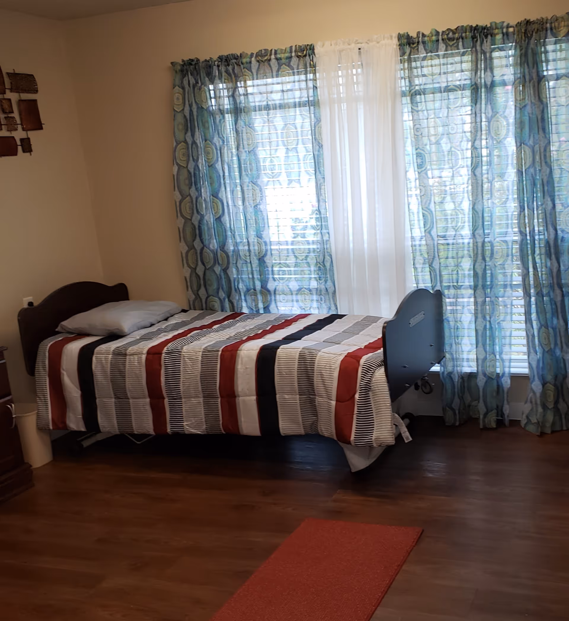 A bedroom with a single bed covered with a striped comforter in shades of red, black, gray, and white. The bed has a gray pillow and is positioned against a wall with a window covered by blue patterned curtains and a sheer white curtain in the center. The floor is wooden, and there is a red rug partially visible in the foreground.