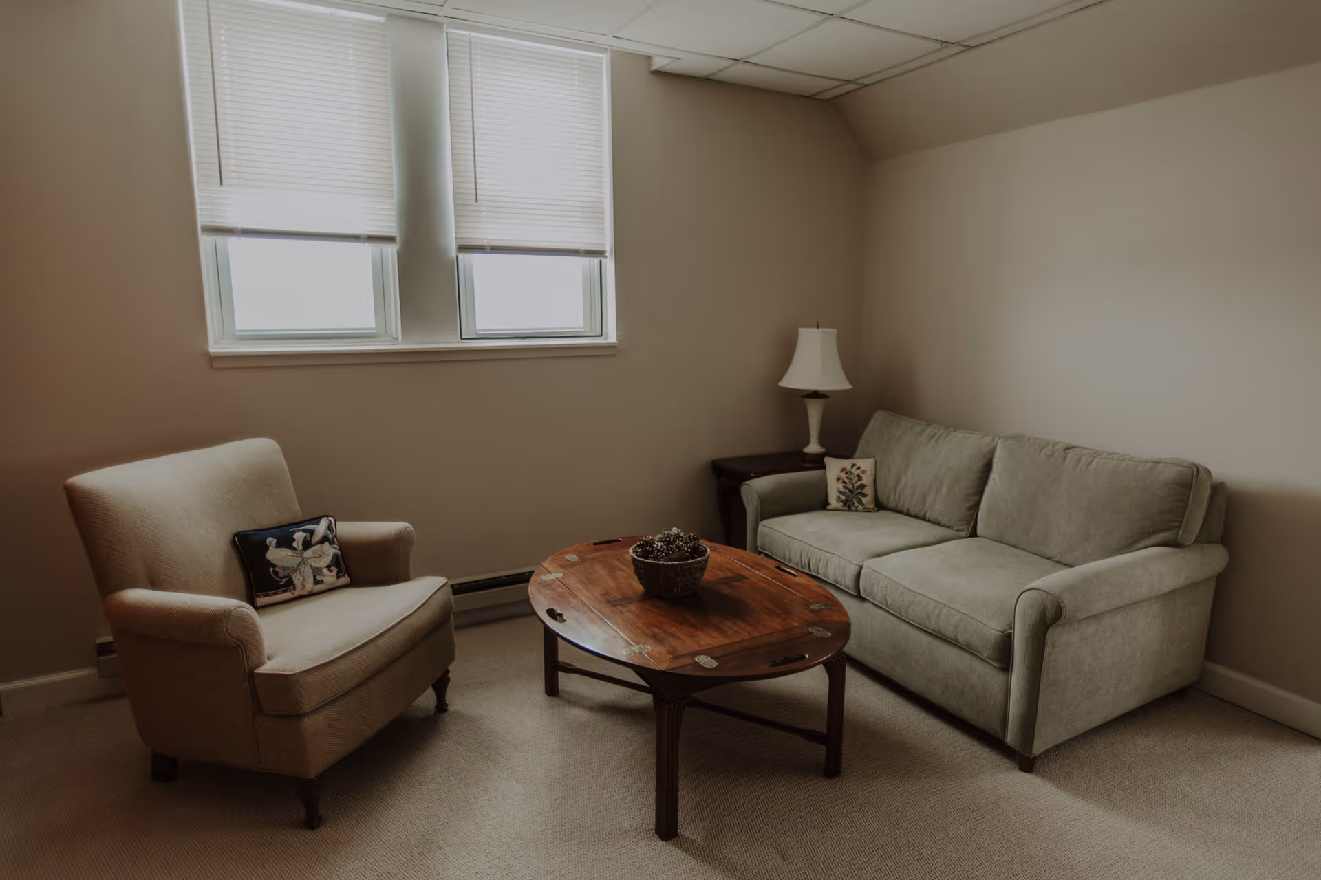 A small living room with a sofa, armchair, wooden coffee table and lamp beneath two windows.
