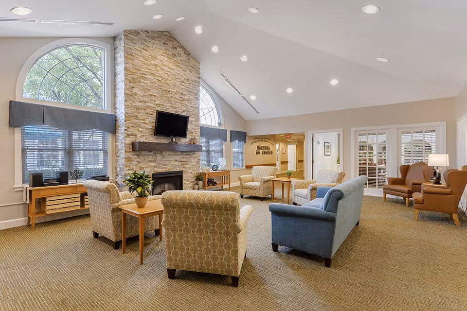 Spacious senior living lounge with multiple armchairs and sofas arranged around a stone fireplace and wall-mounted TV beneath large arched windows.