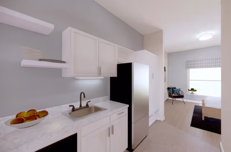 A bright studio interior featuring a small kitchen with white cabinets, marble countertop, and stainless steel refrigerator opening into a bedroom area with a bed and chair.