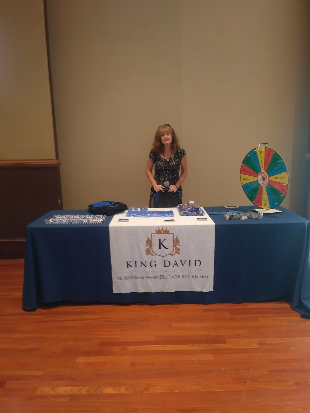A woman standing behind a table covered with a blue tablecloth and a white banner that reads 'King David Nursing & Rehabilitation Center.' The table has various promotional items and a colorful prize wheel on the right side. The setting appears to be indoors with wooden flooring and plain walls.