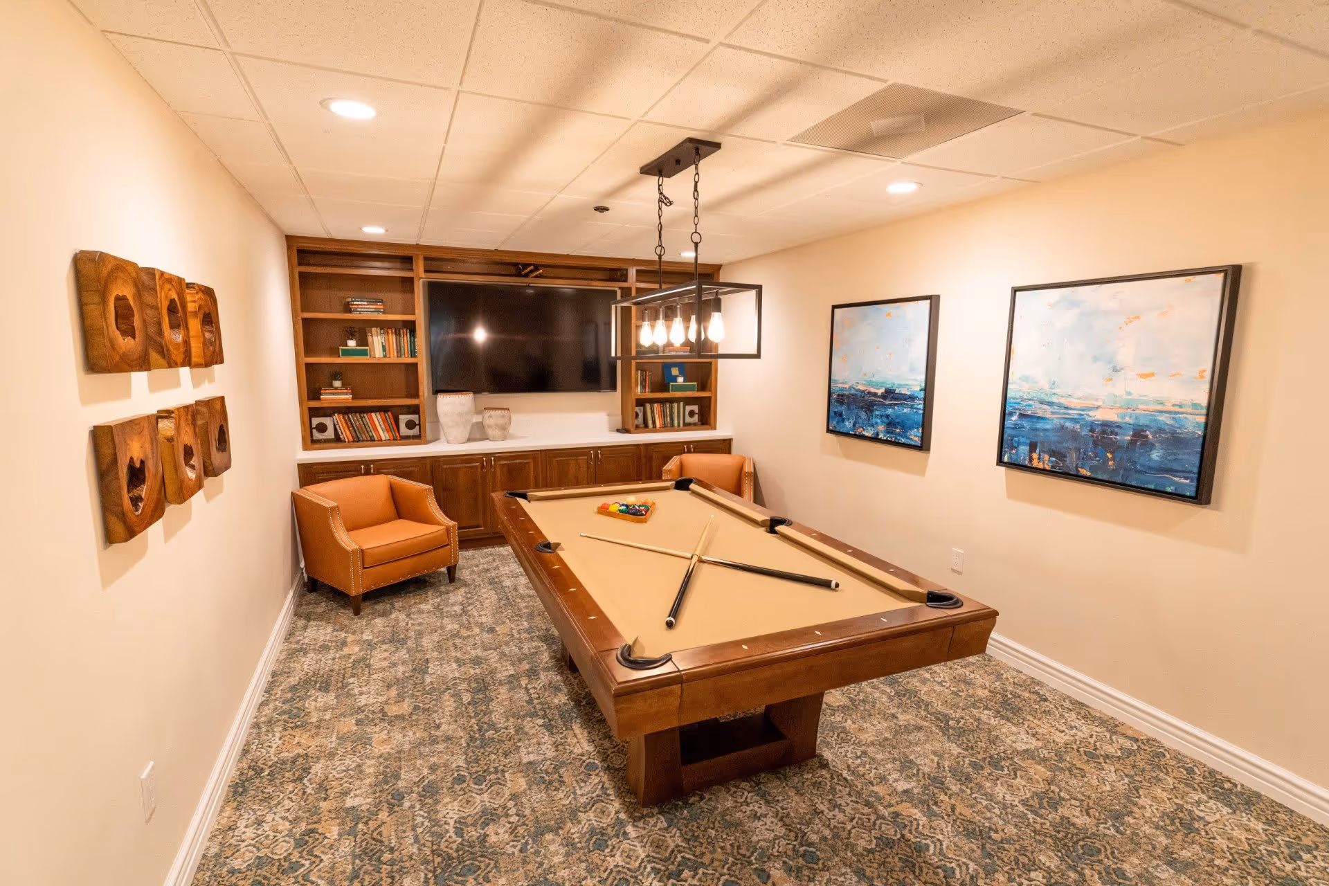 A cozy game room with a pool table in the center, two pool cues crossed on the table, and a rack of billiard balls. The room features a patterned carpet, beige walls with two abstract paintings, a built-in wooden bookshelf with a large flat-screen TV, and two brown leather armchairs. A modern rectangular light fixture hangs above the pool table.