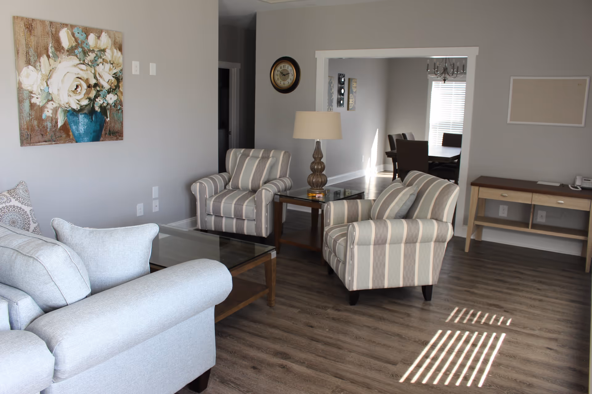 Bright living room with striped armchairs, a sofa, glass coffee table, lamp, and a view into a dining area.
