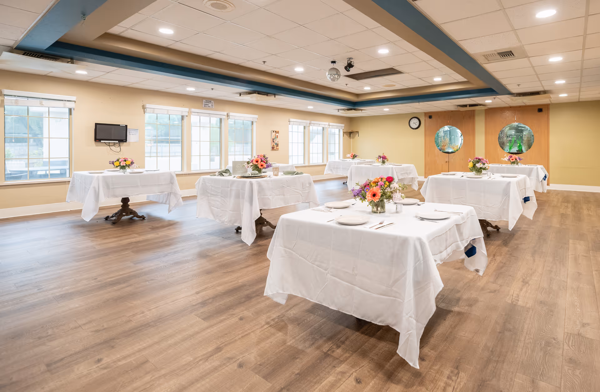 A spacious dining room with six tables covered in white tablecloths, each set with plates, utensils, and floral centerpieces. The room has large windows letting in natural light, wood flooring, and a ceiling with recessed lighting and a disco ball.