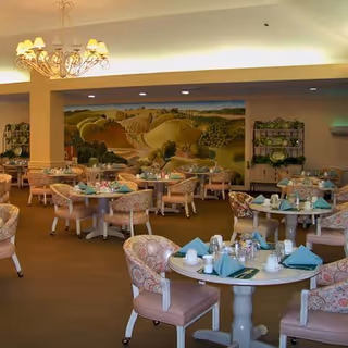 A dining room with multiple round tables set with cups, plates, and folded napkins. The chairs have floral upholstery and the room features a large mural of a countryside landscape on the far wall. There is a chandelier hanging from the ceiling and shelves with decorative items along the walls.