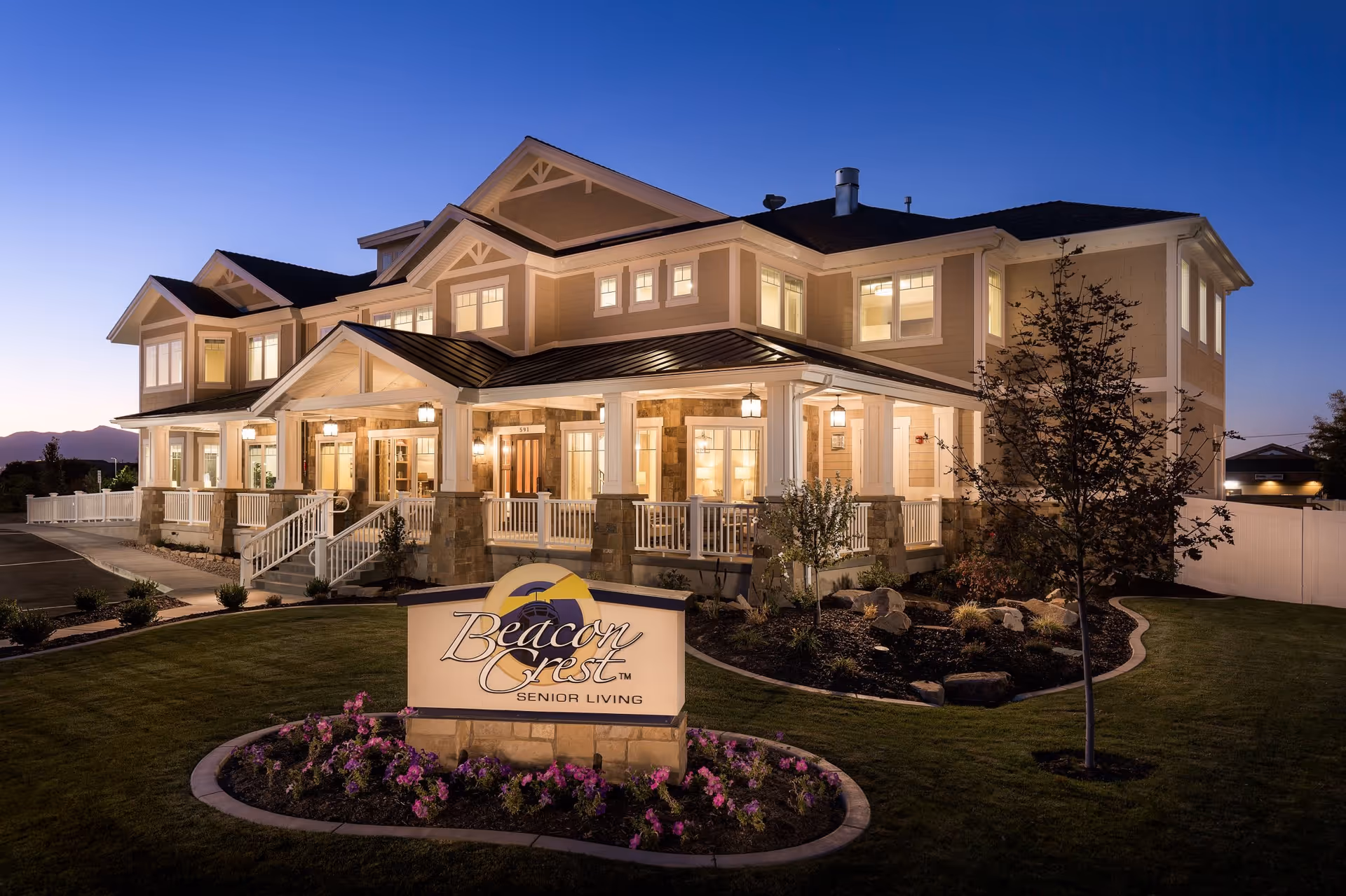 Exterior view of Beacon Crest Senior Living Draper building at dusk, showing a well-lit two-story structure with a porch, landscaped garden with flowers, and a sign in front displaying the facility's name.