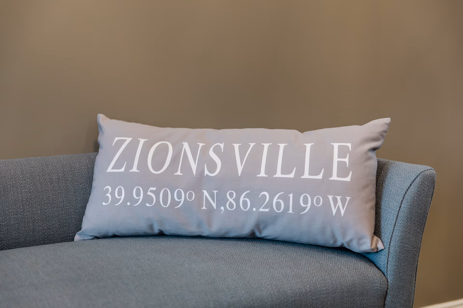 A decorative pillow reading "ZIONSVILLE" with coordinates sits on a gray upholstered chair against a beige wall.