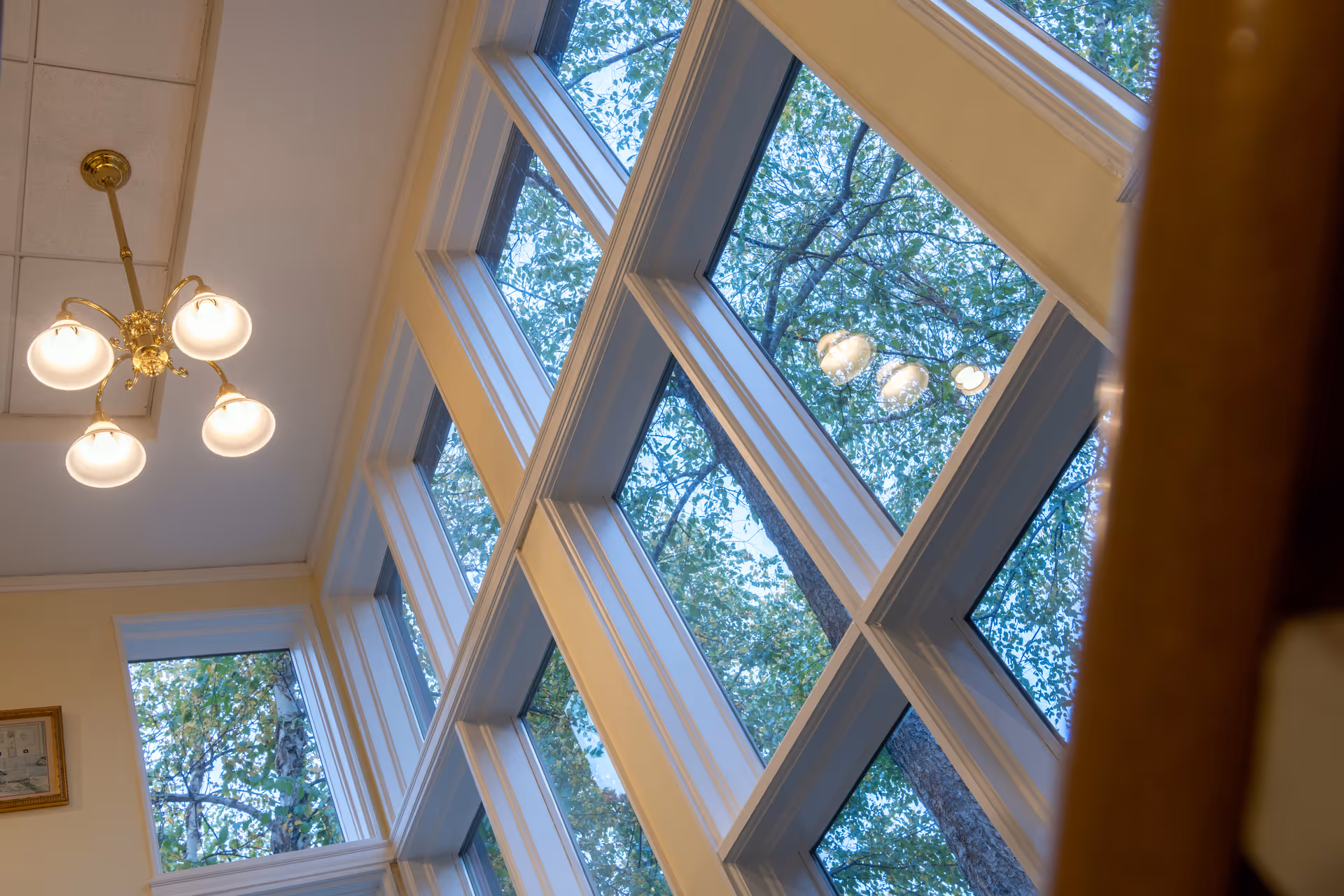 Interior view of tall multi-pane windows with trees visible outside and a brass ceiling chandelier.