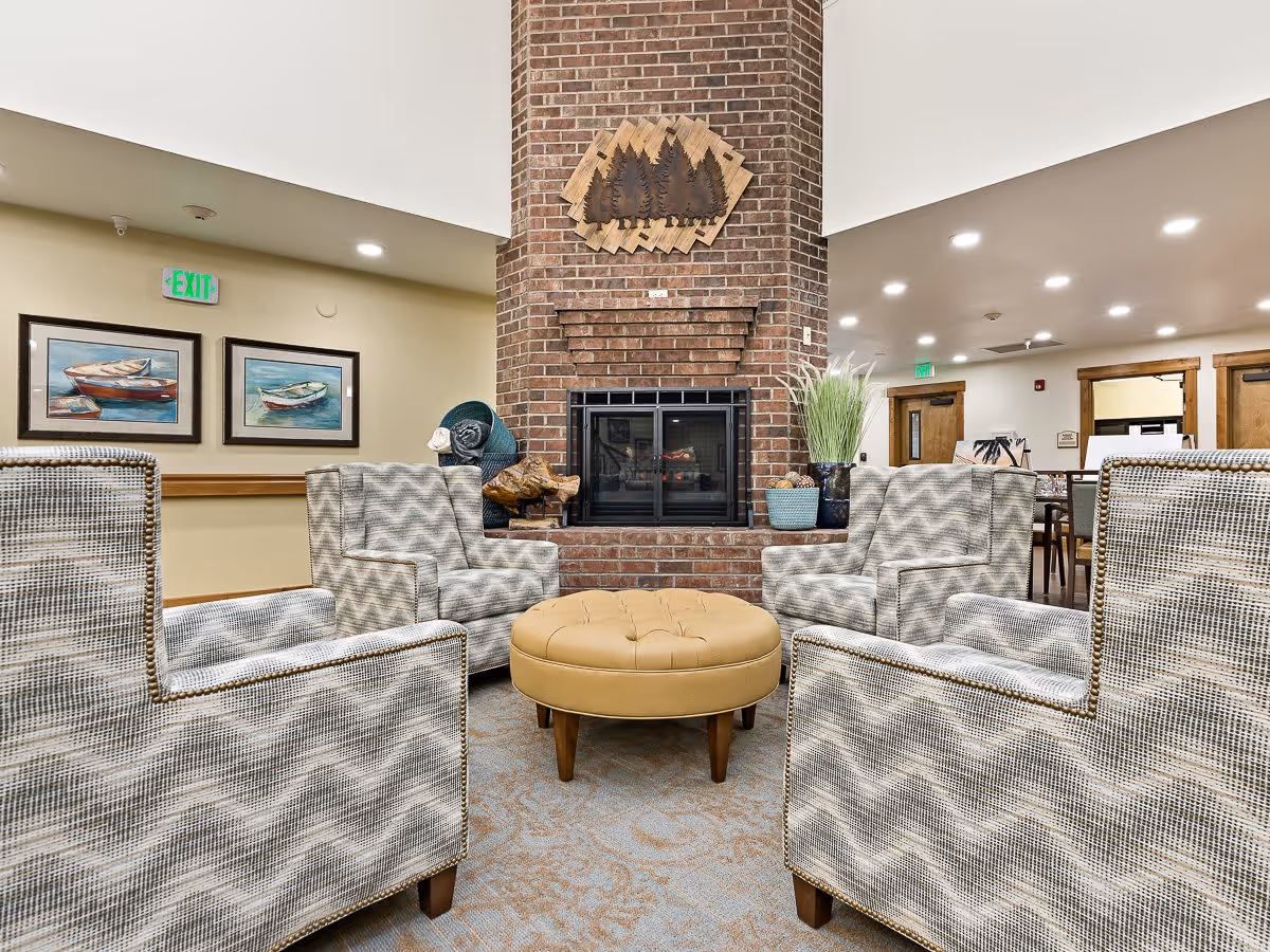 A cozy seating area in a memory care facility featuring four patterned armchairs arranged around a round tan ottoman in front of a brick fireplace. The fireplace has a decorative wooden wall art above it depicting trees. The room has soft lighting, framed paintings of boats on the wall, and some decorative plants and items on the fireplace hearth.