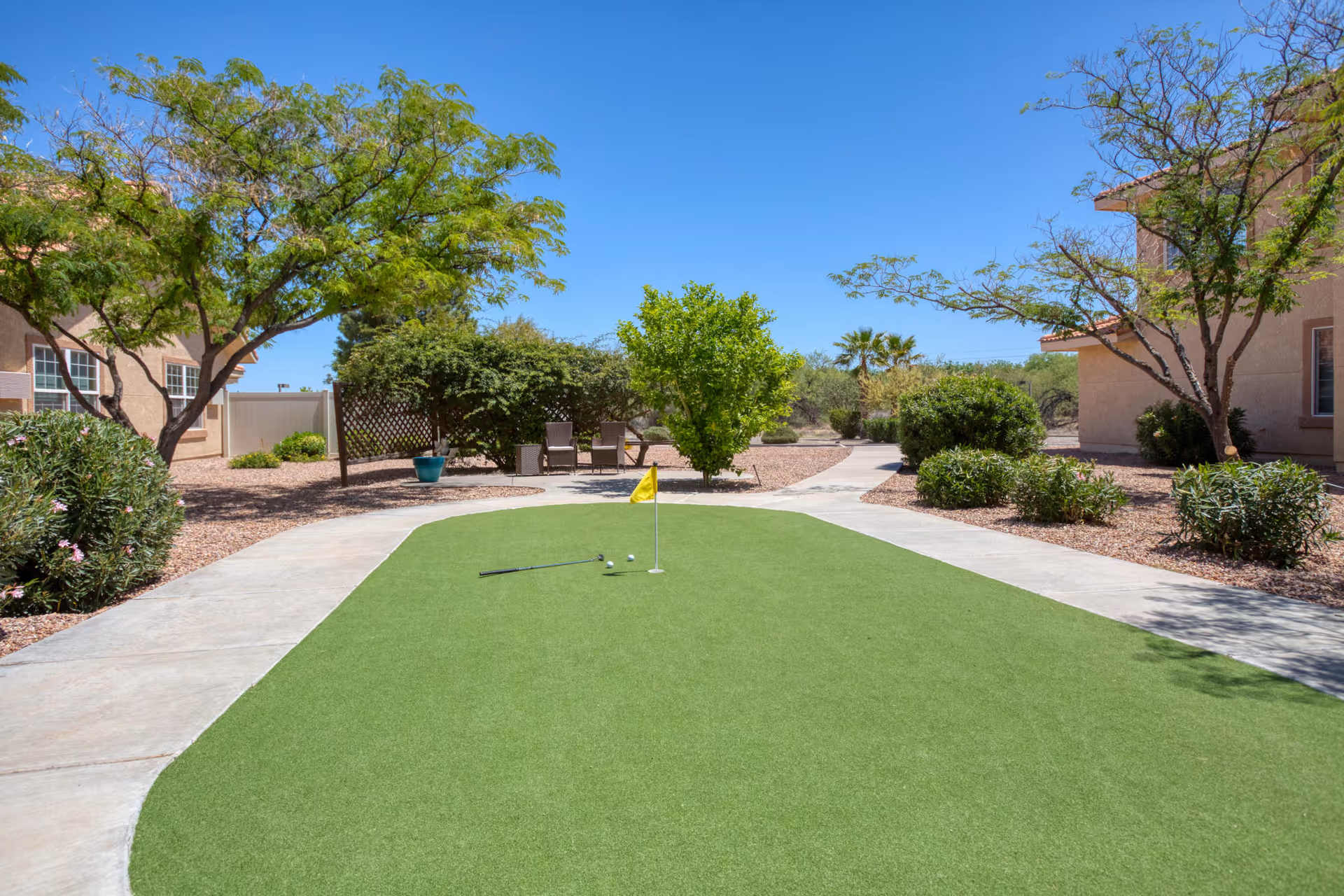 A courtyard putting green with a small flag and golf balls surrounded by walkways, desert landscaping, trees and residential buildings under a clear blue sky.