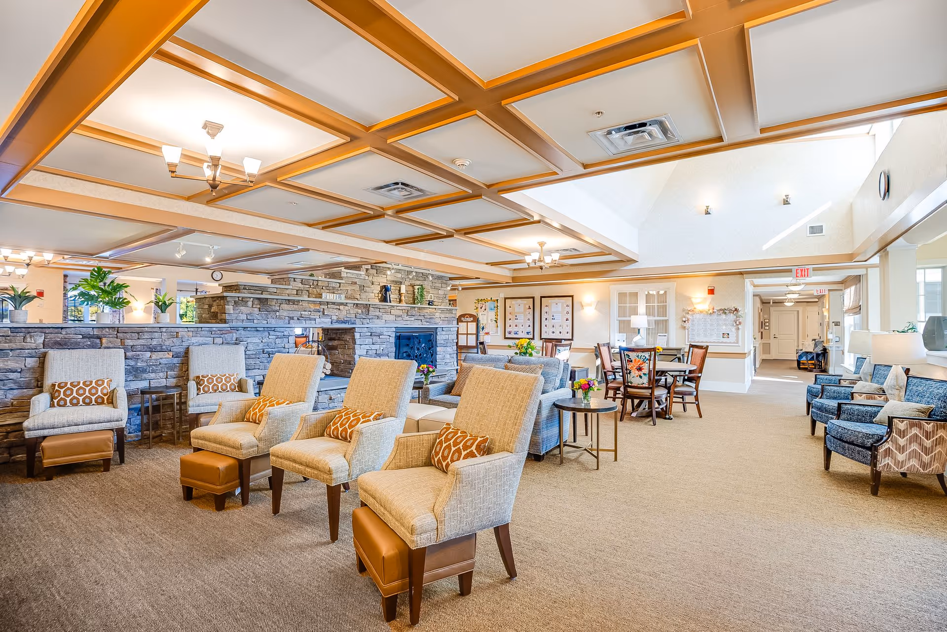 A spacious and well-lit senior living facility common area with comfortable armchairs featuring patterned cushions, small side tables, and a stone fireplace in the center. The ceiling has a decorative wooden grid design with multiple light fixtures. In the background, there are dining tables and chairs, bulletin boards on the walls, and large windows allowing natural light to fill the room.
