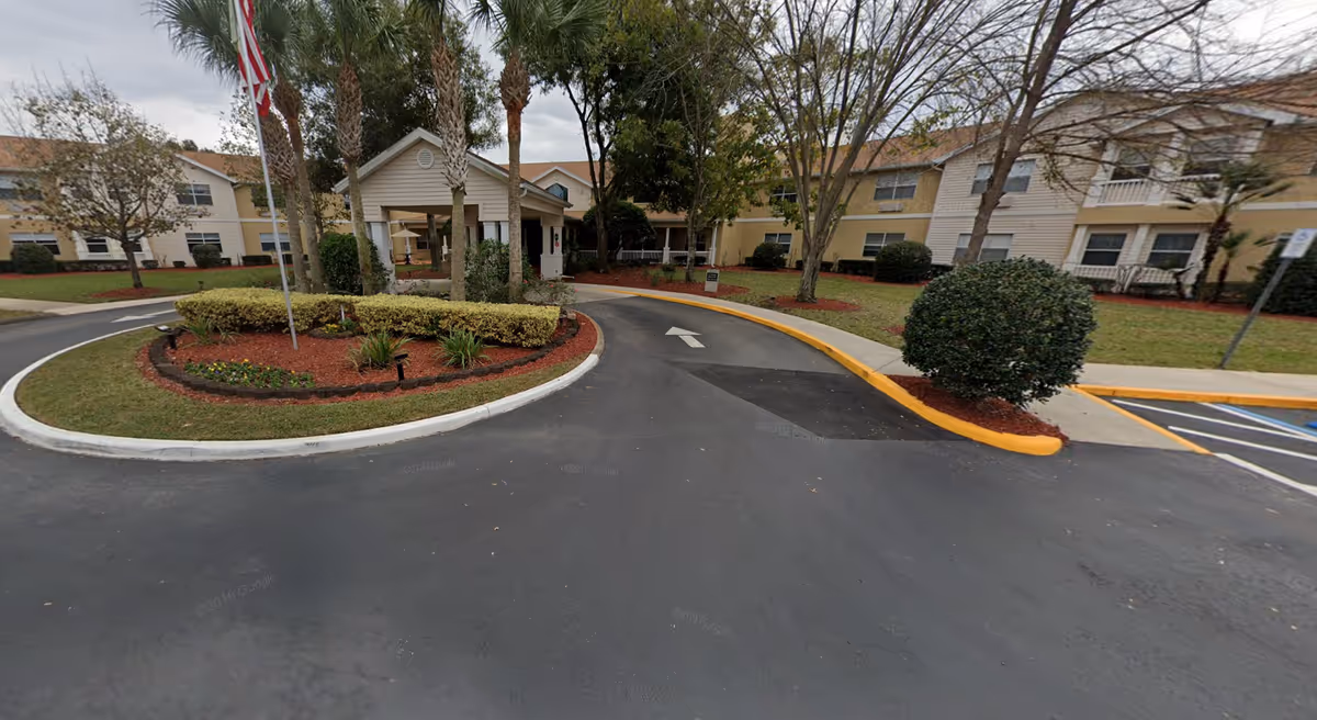 Exterior view of Ansley Parke at Orange City senior living facility showing a circular driveway with landscaped center island featuring palm trees and bushes, an American flag, and the entrance to the building with beige walls and a covered porch.