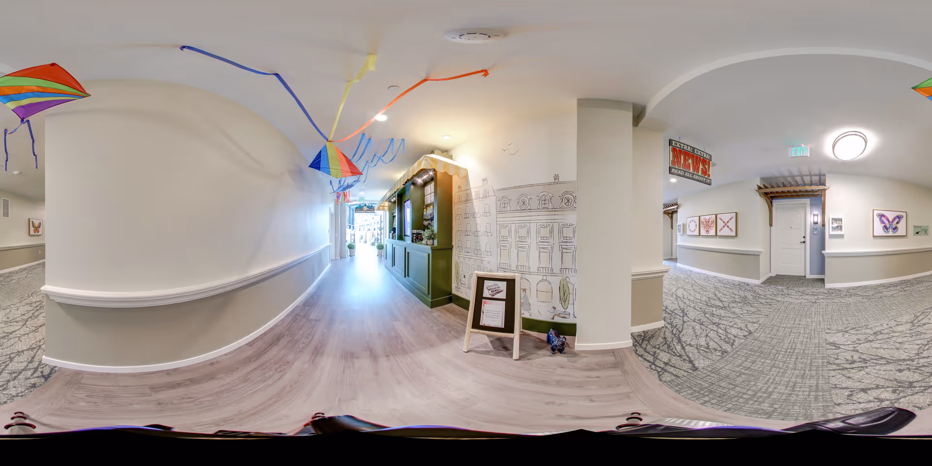 A wide hallway in a senior living facility with light-colored walls and flooring. Colorful kites hang from the ceiling along the corridor. On the right side, there is a green reception or service counter with a small chalkboard sign in front of it. The walls have decorative artwork, including framed pictures and a mural sketch. The hallway leads to a bright exit door at the far end.
