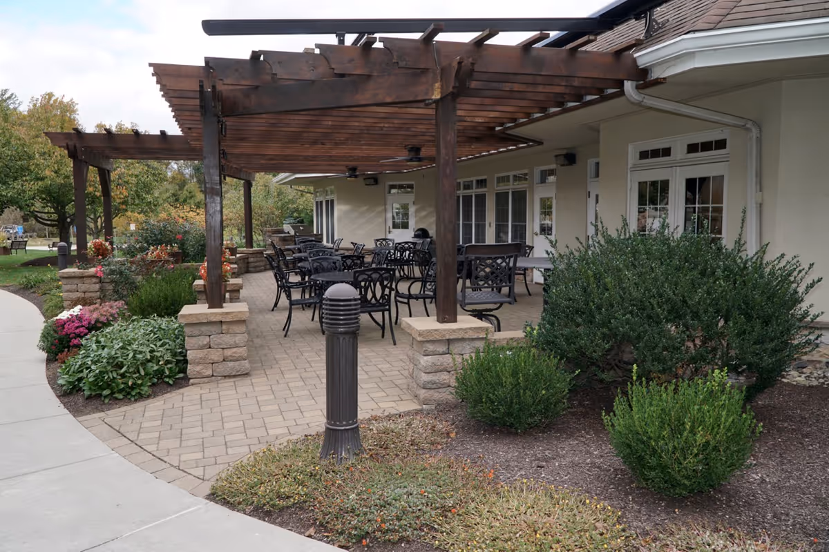 Outdoor patio area with a wooden pergola covering several black metal tables and chairs. The patio is paved with bricks and surrounded by landscaped bushes and flowers. The building adjacent to the patio has multiple windows and doors with white frames.