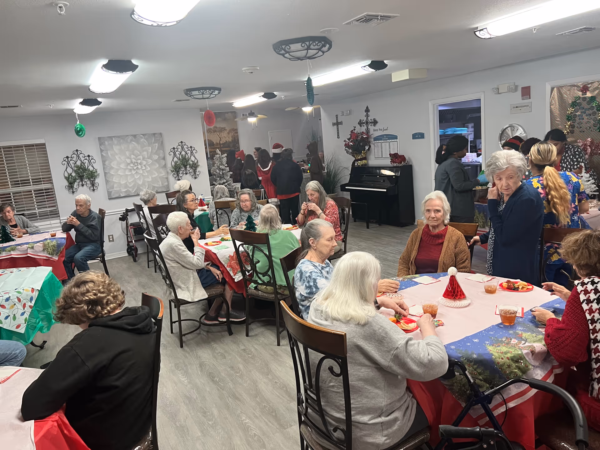 A group of elderly people seated around tables in a decorated room, enjoying food and drinks. The room has festive holiday decorations including a small Christmas tree, hanging ornaments, and tablecloths with holiday themes. Several caregivers or staff members are standing and interacting with the residents. The environment appears warm and social.