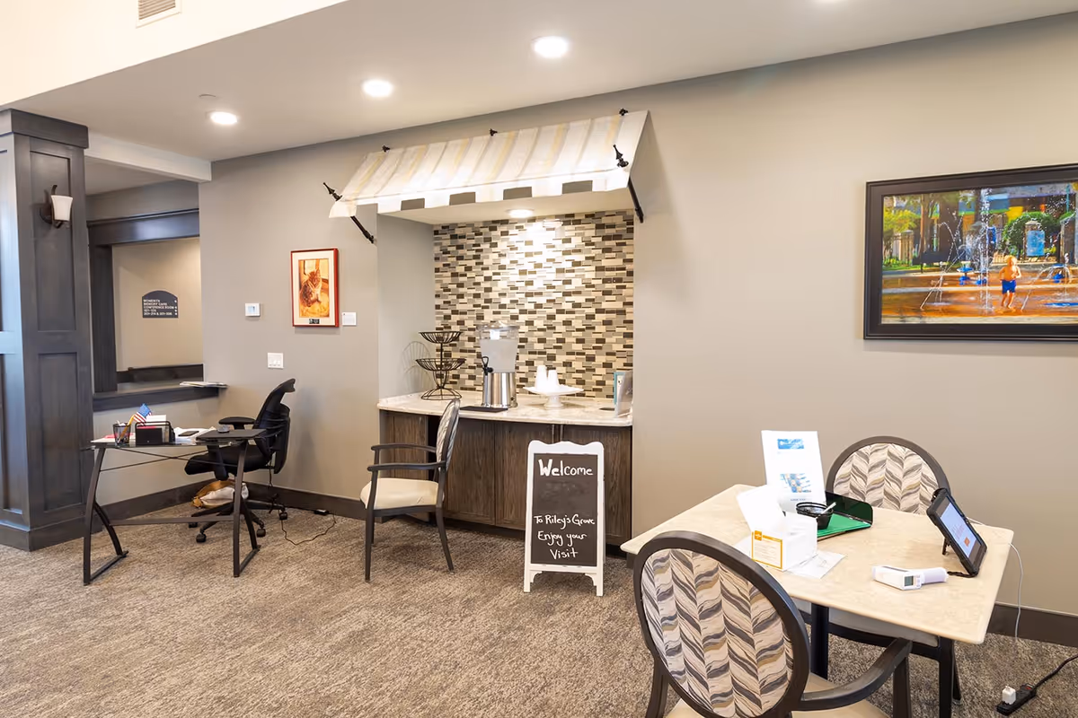 Interior view of a reception or common area in Riley's Grove Assisted Living & Memory Care featuring a small table with two chairs, a desk with an office chair, a refreshment station with a water dispenser and cups, a welcome sign, and a framed painting on the wall.