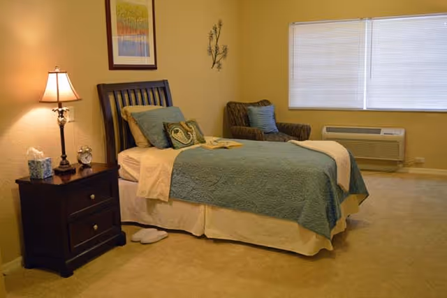 A cozy bedroom with a single bed covered in a blue quilt and beige sheets, a wooden nightstand with a lamp, alarm clock, and tissue box, a cushioned armchair with a blue pillow, a large window with closed blinds, and a beige carpeted floor.