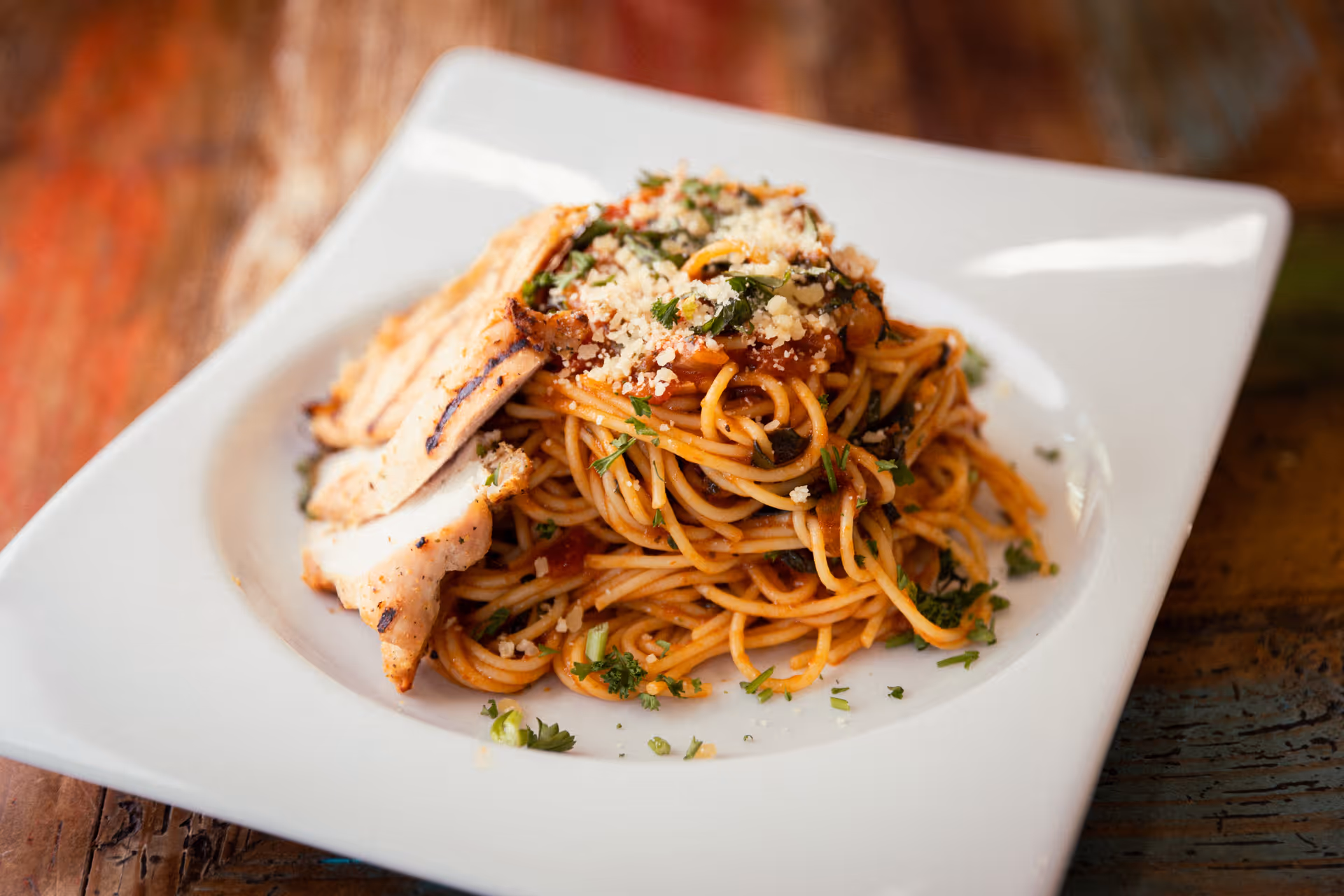 A plate of spaghetti topped with grilled chicken slices and grated cheese on a white square plate.