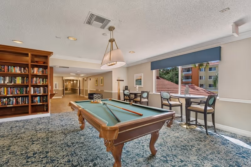 A well-lit indoor recreational room featuring a pool table with cues and balls arranged on it. To the right, there are two small round tables each with two chairs, positioned near large windows with a blue valance, offering a view of nearby buildings and greenery. On the left side, there is a wooden bookshelf filled with books. The room has a patterned carpet and a hanging light fixture above the pool table.