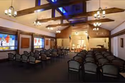 Interior chapel-style meeting room with rows of chairs facing an altar, exposed wooden ceiling beams, stained glass windows and chandeliers.