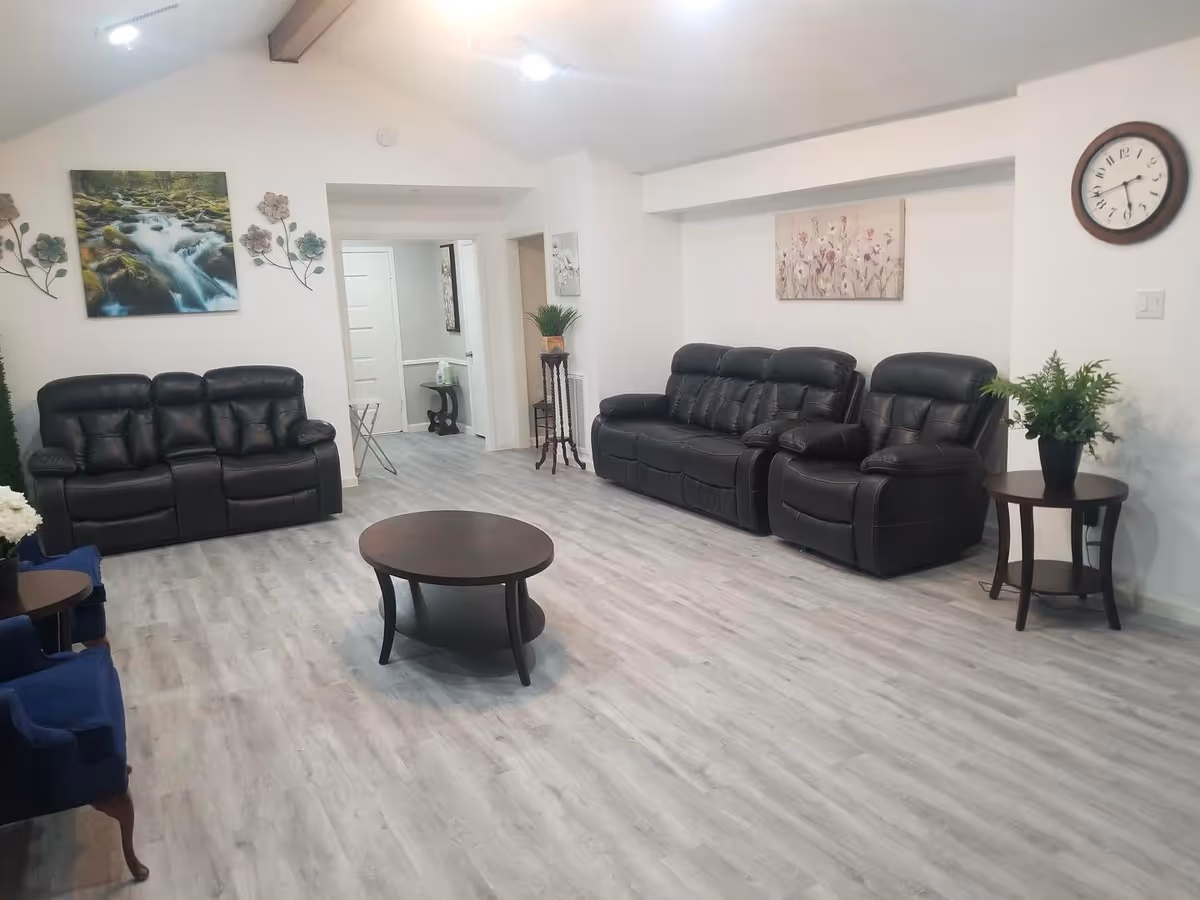 Bright senior living lounge with black leather seating, plants, wall clock, and artwork.