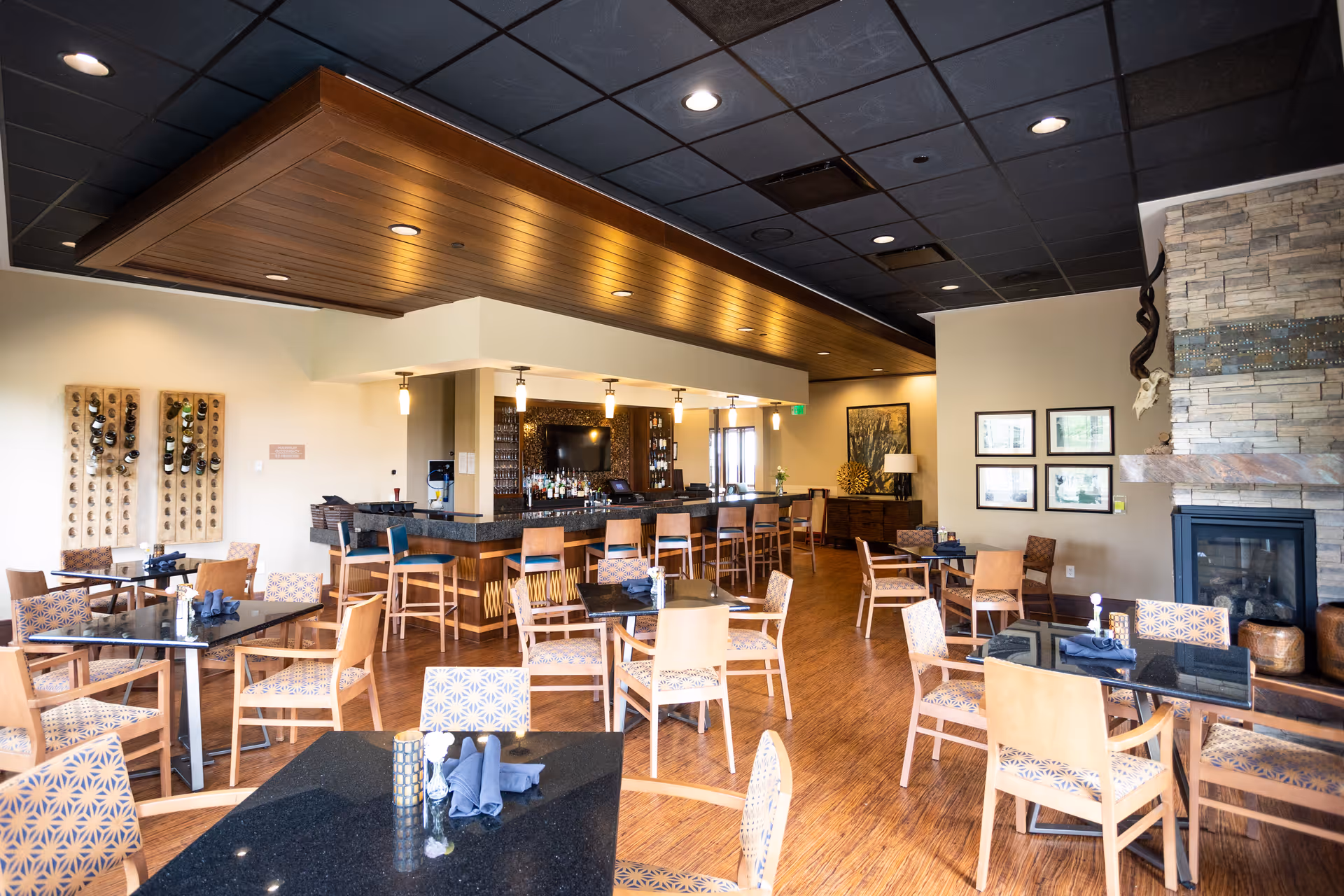 A spacious dining area with multiple tables and chairs arranged neatly. The room features a bar with high stools, a stone fireplace on the right, and decorative wall art. The ceiling has recessed lighting and a wooden accent above the bar area.