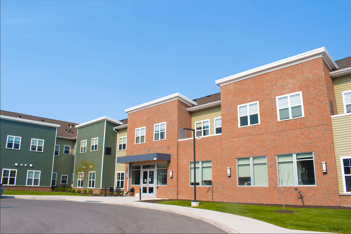 Front exterior of a modern multi-story brick and green-siding senior living building with an entrance canopy and windows under a clear blue sky.