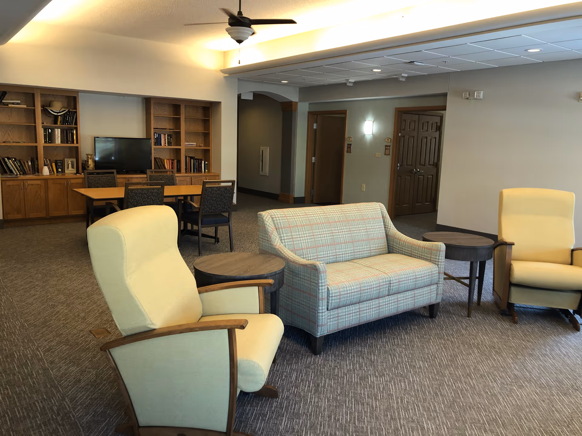 A cozy common area in a senior living facility with two yellow armchairs, a plaid loveseat, and two round wooden side tables. In the background, there is a wooden bookshelf with books and a flat-screen TV, along with a table and chairs. The room has carpeted flooring, neutral-colored walls, and ceiling lights.