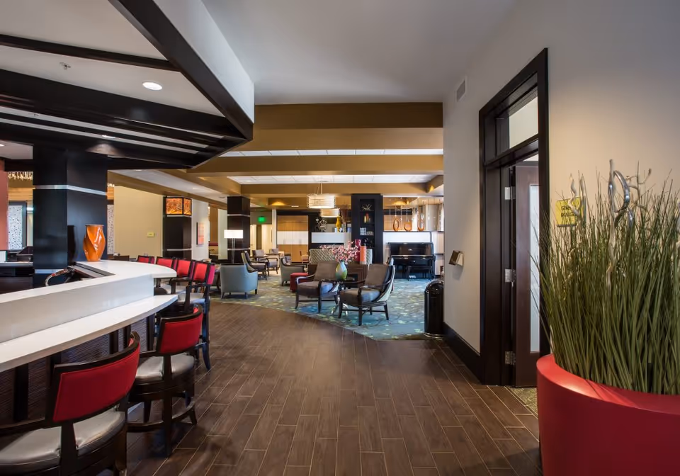 Interior view of a senior living facility lounge area with a bar counter on the left featuring red cushioned bar stools. The space has wooden flooring, modern furniture including armchairs and tables, decorative plants, and a piano in the background. The ceiling has exposed beams and pendant lighting.